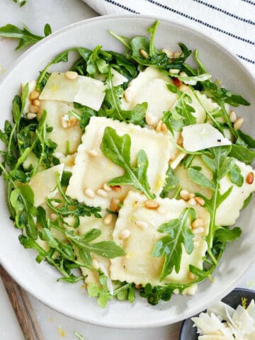 Ravioli pasta salad with arugula in a bowl surrounded by napkin, lemon, spoon, and cheese.
