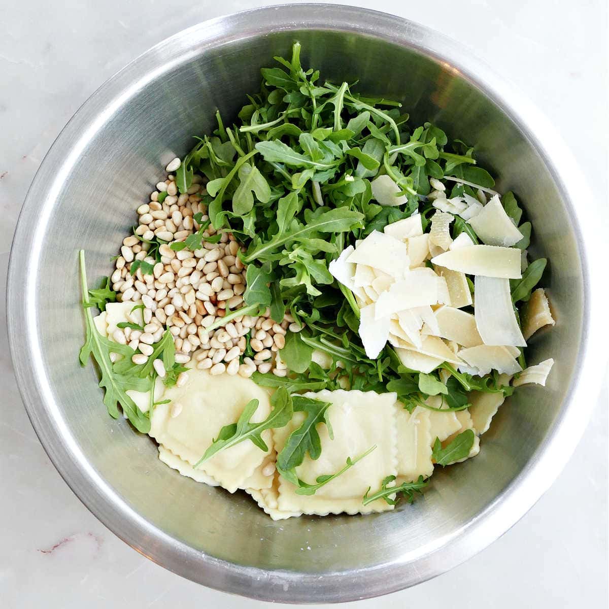 Ravioli, pine nuts, arugula, and shaved parmesan cheese in a mixing bowl.