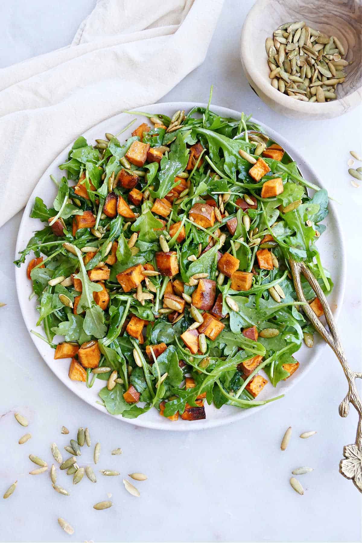 A plate of arugula sweet potato salad with cumin lime dressing on top next to a bowl of sunflower seeds.