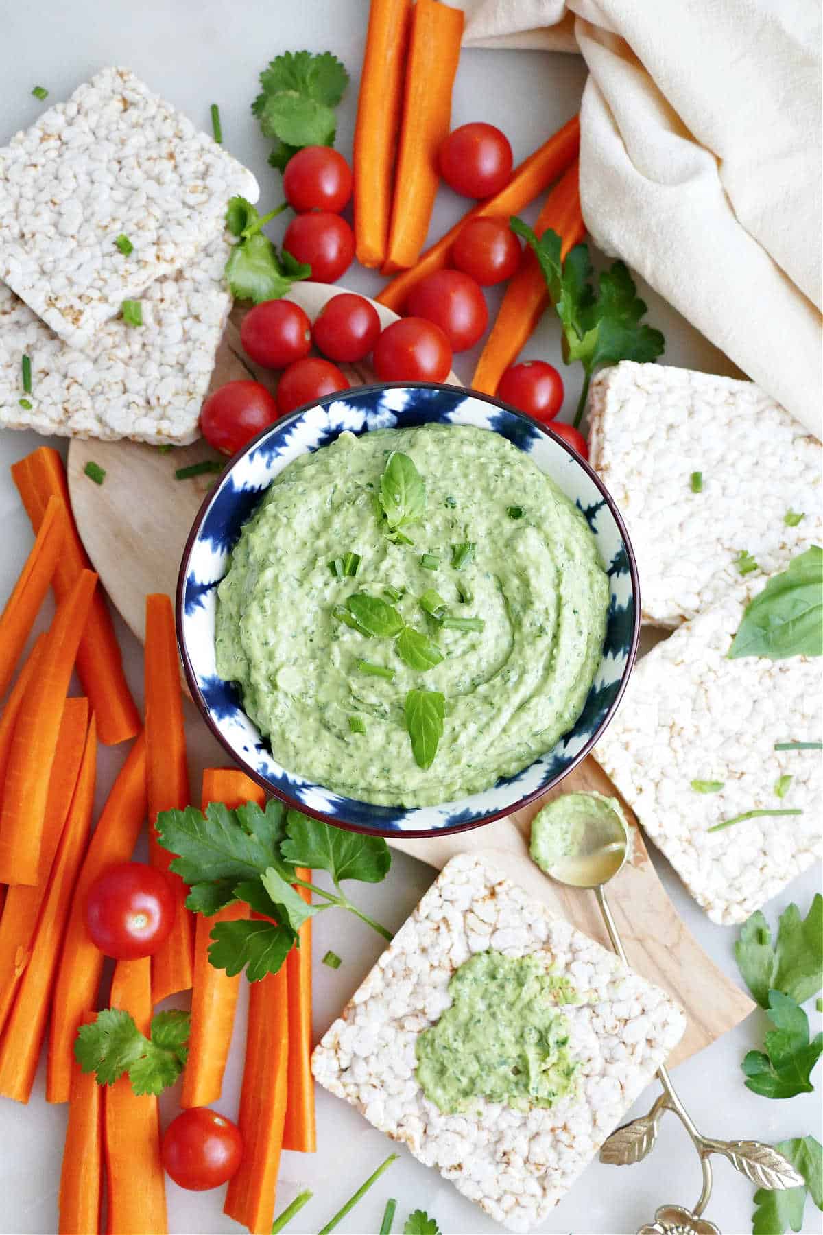 Avocado green goddess dressing in a small bowl surrounded by vegetables and rice cakes.