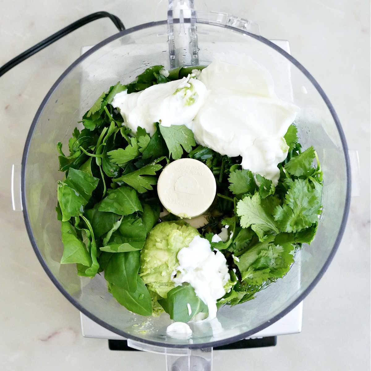 Fresh herbs, Greek yogurt, avocado, and seasonings in a food processor.
