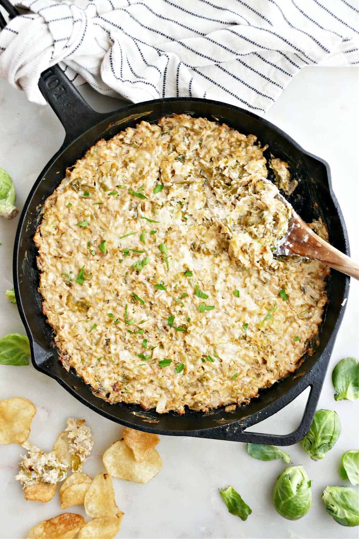 Brussels Sprouts Dip in a cast iron pan with a wood spoon next to chips and fresh Brussels sprouts.
