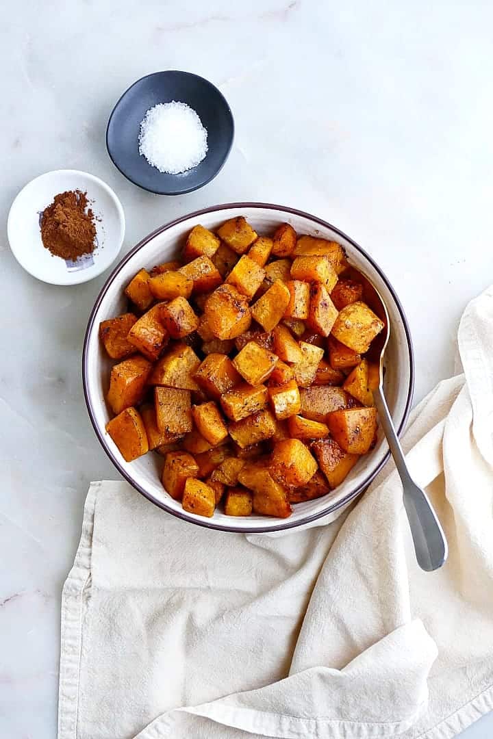 A bowl full of roasted cinnamon butternut squash cubes next to two bowls of cinnamon and sugar.