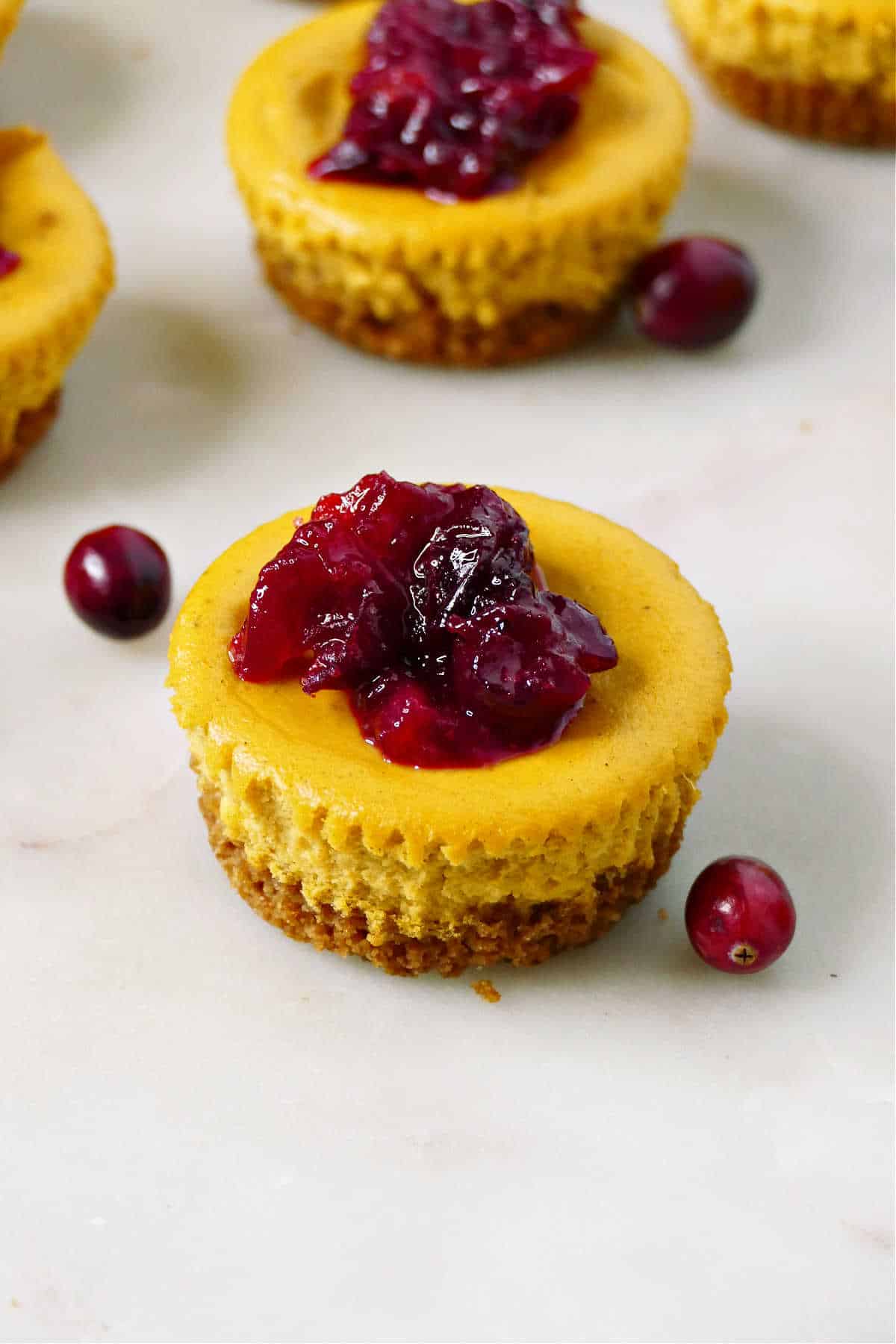 A close up of a mini pumpkin cranberry cheesecake next to two fresh cranberries. 