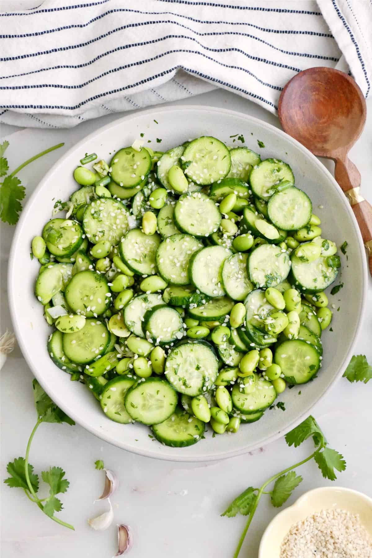 Cucumber edamame salad in a serving bowl next to garnishes, a napkin, and a spoon.