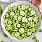 Cucumber edamame salad in a serving bowl next to garnishes, a napkin, and a spoon.