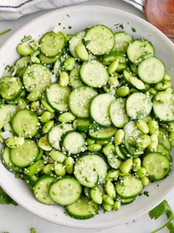 Cucumber edamame salad in a serving bowl next to garnishes, a napkin, and a spoon.