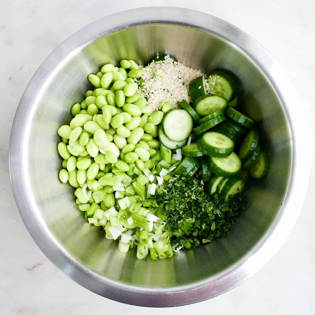 Edamame, sesame seeds, sliced cucumber, cilantro, and green onions in a mixing bowl.