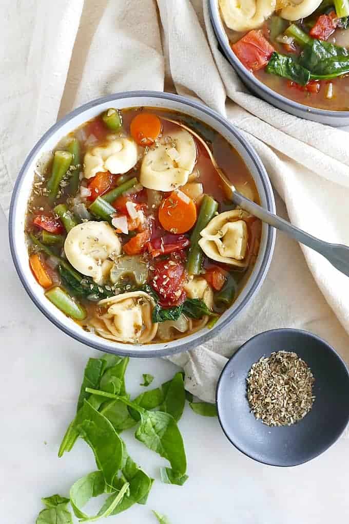 Vegetable tortellini soup in two bowls with a spoon next to towel and seasoning. 