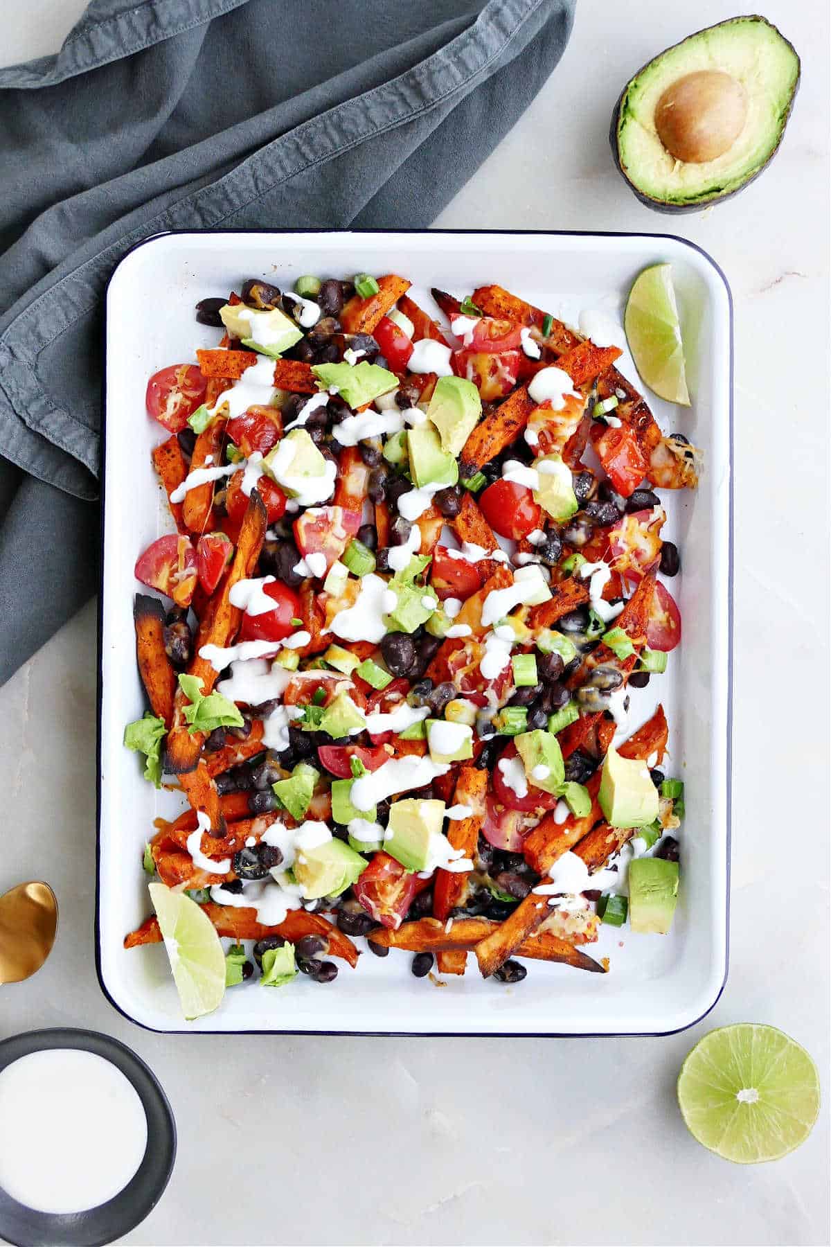 A plate of loaded sweet potato fries next to half of an avocado and gray kitchen towel.