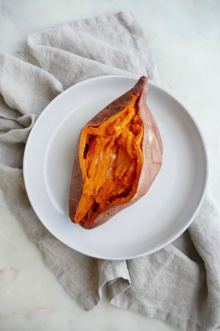 A microwaved sweet potato on a white plate sitting on top of a gray dish cloth.