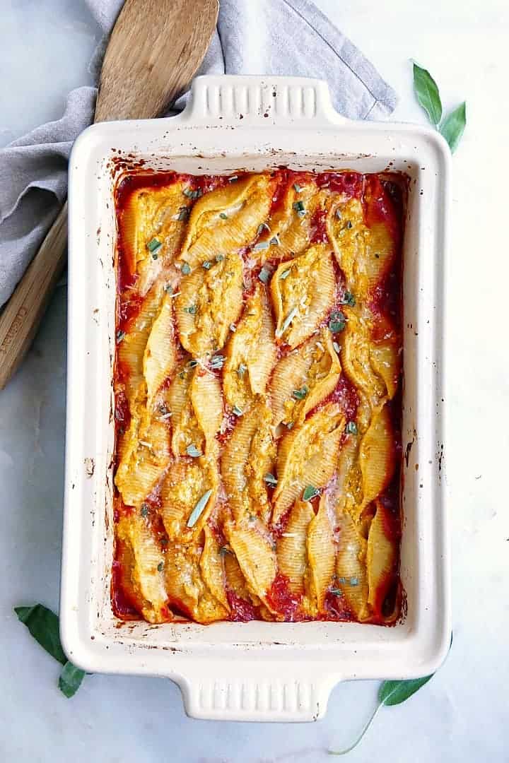 White baking dish filled with ricotta pumpkin stuffed shells next to a wooden spoon and a grey dish towel. 