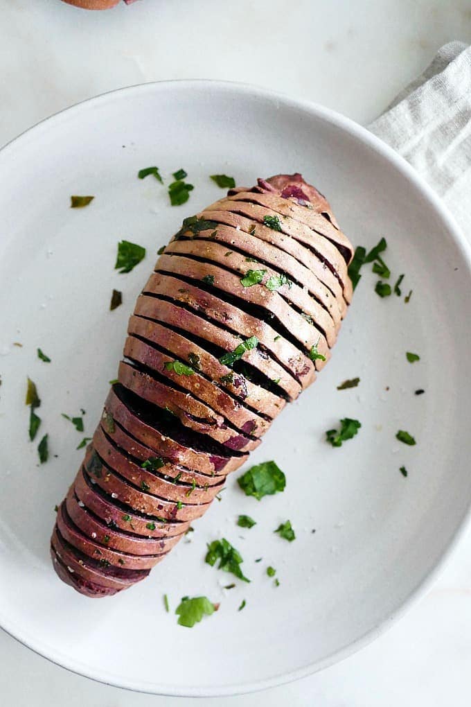 Hasselback purple sweet potato on a white plate sprinkled with parsley.