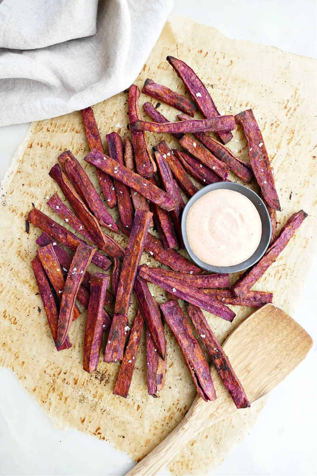 Parchment paper with purple sweet potato fries on top of it next to a cup of ketchup and Greek yogurt dipping sauce.