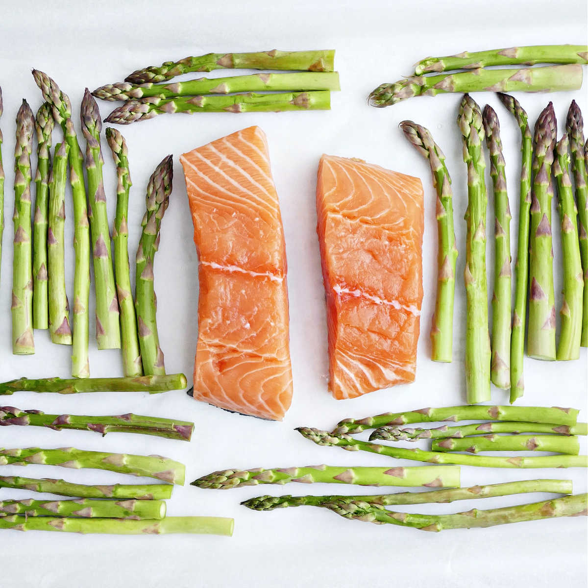 Raw salmon fillets in the center of a lined baking sheet surrounded by asparagus.