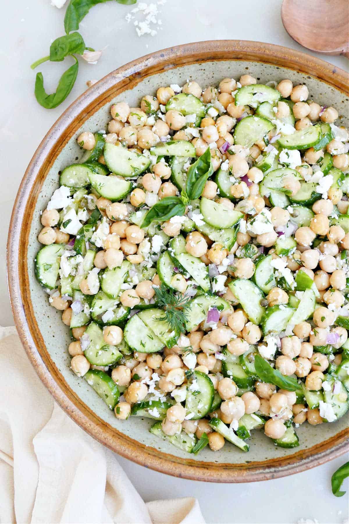 Cucumber, chickpeas, feta, and herbs with dressing in a mixing bowl.