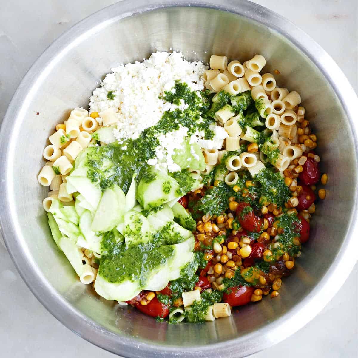 Pasta, cooked tomatoes and corn, cucumber, feta cheese, and basil vinaigrette in a mixing bowl.