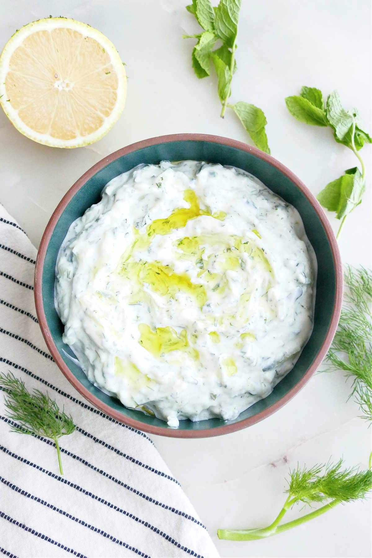 Cucumber yogurt sauce with olive oil in a bowl surrounded by herbs, lemon, and a napkin.