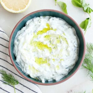 Cucumber yogurt sauce with olive oil in a bowl surrounded by herbs, lemon, and a napkin.
