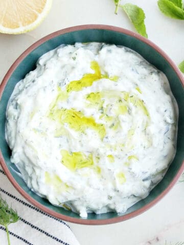 Cucumber yogurt sauce with olive oil in a bowl surrounded by herbs, lemon, and a napkin.