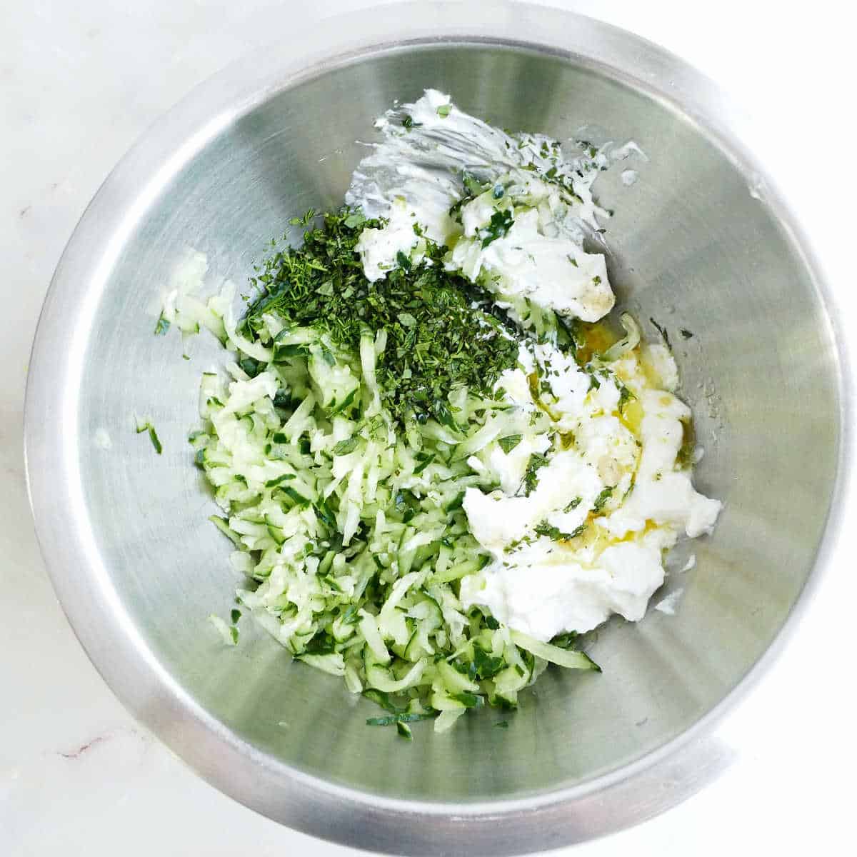 Grated cucumber, chopped herbs, olive oil, lemon juice, seasonings, and yogurt in a mixing bowl.