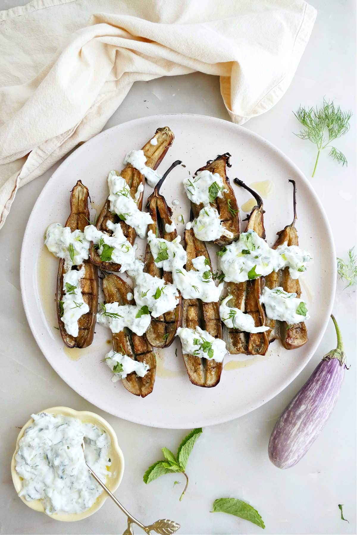Baby eggplants with cucumber sauce on a plate next to napkins and ingredients.