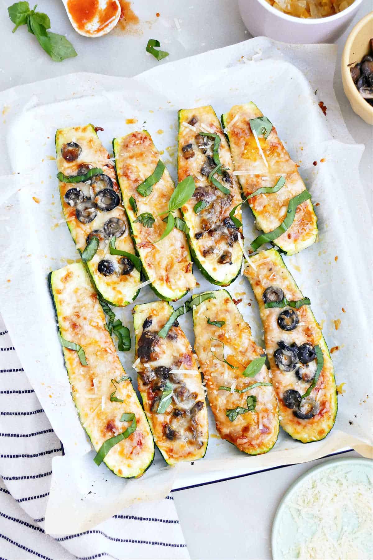 Eight zucchini pizza boats on a rectangular serving dish surrounded by toppings.