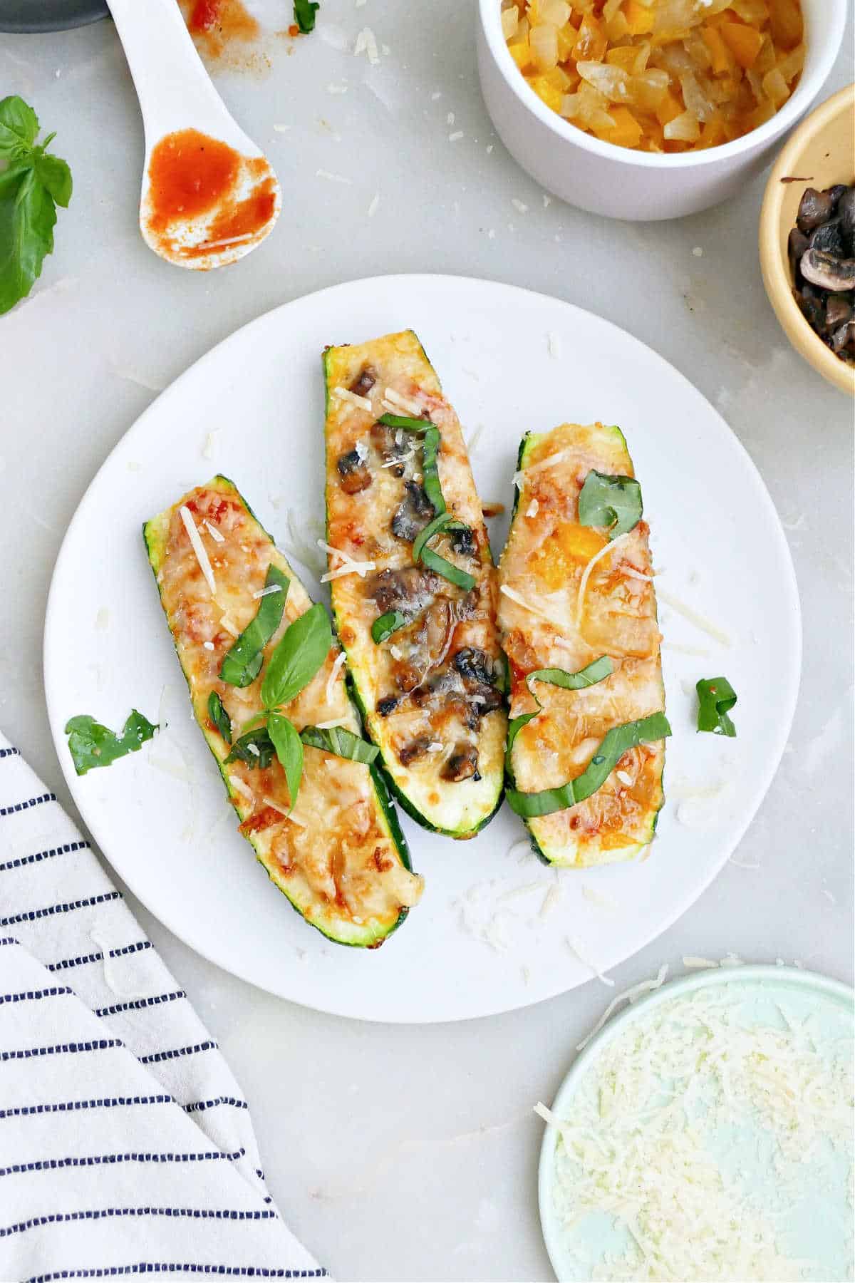 Three zucchini pizza boats on a plate next to toppings and a napkin.