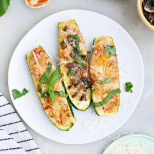 Three zucchini pizza boats on a plate next to toppings and a napkin.