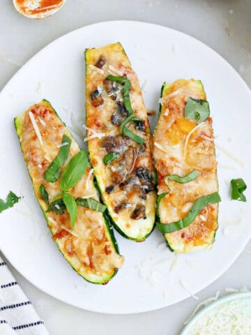 Three zucchini pizza boats on a plate next to toppings and a napkin.