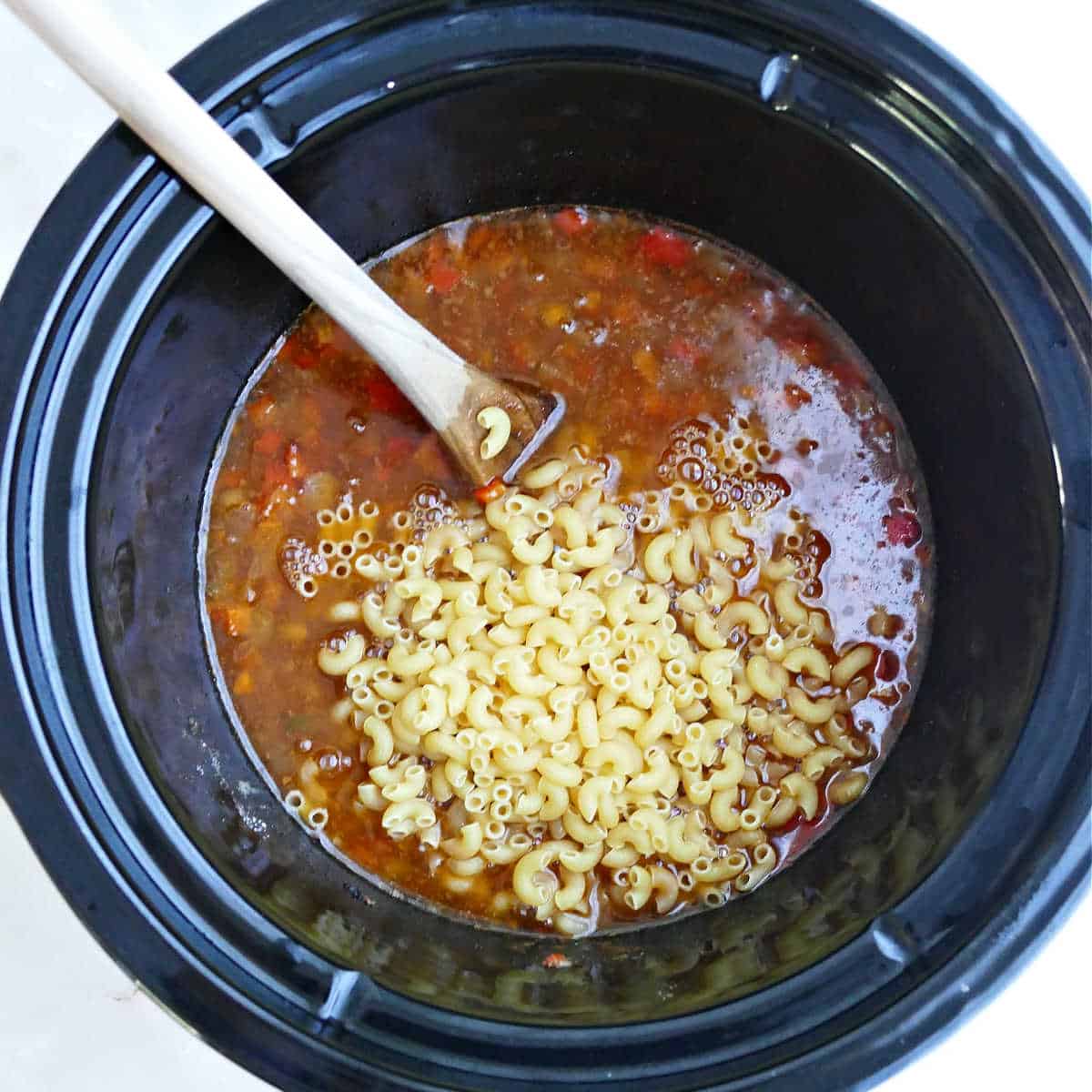 Dried macaroni being stirred into slow cooker hamburger helper.