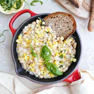 White beans, corn, basil, and bread in a red skillet next to ingredients.