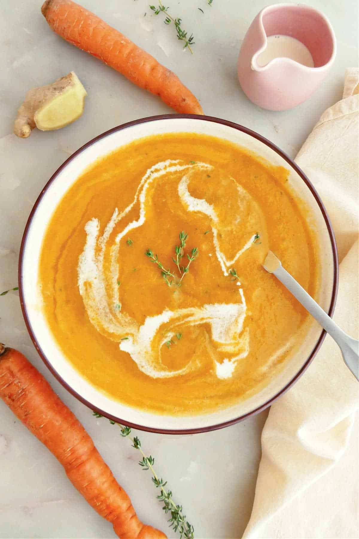 Carrot ginger soup with cream and thyme in a bowl with a spoon surrounded by ingredients.