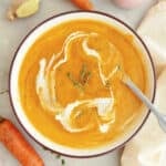 Carrot ginger soup with cream and thyme in a bowl with a spoon surrounded by ingredients.
