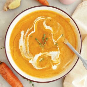 Carrot ginger soup with cream and thyme in a bowl with a spoon surrounded by ingredients.