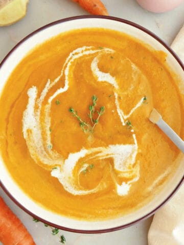 Carrot ginger soup with cream and thyme in a bowl with a spoon surrounded by ingredients.