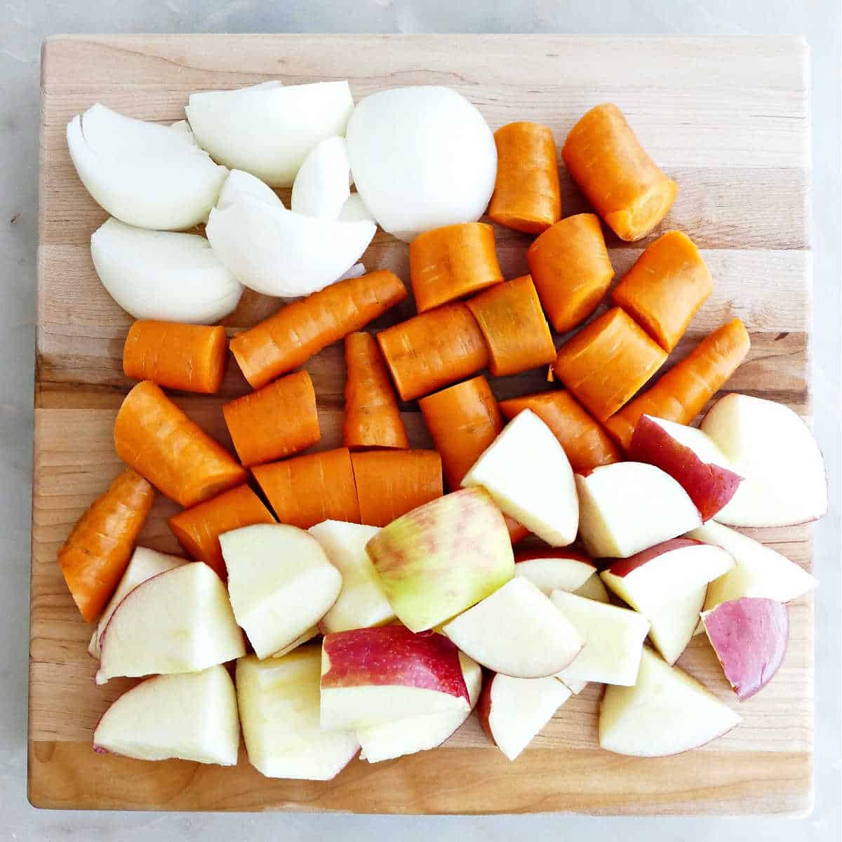 Roughly chopped onion, carrots, and apples on a wood cutting board.