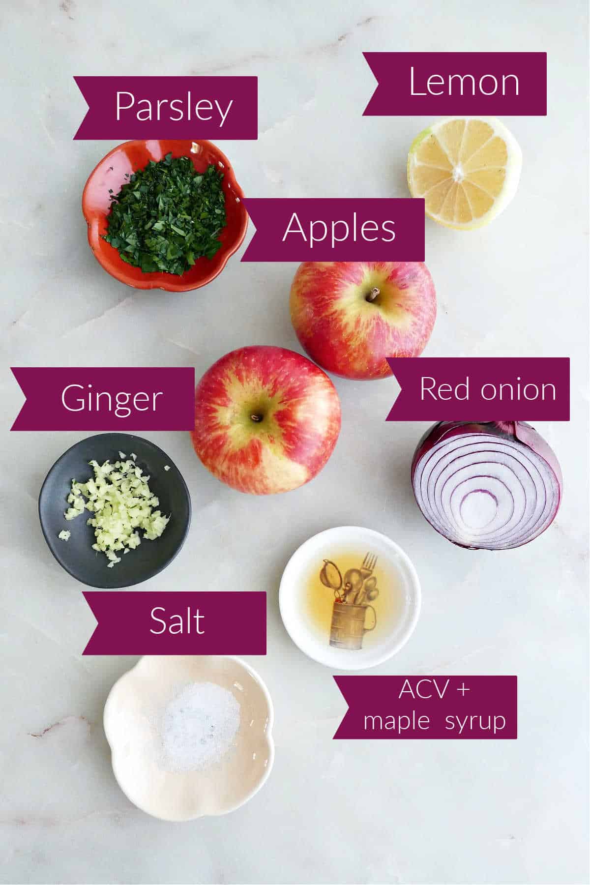 Parsley, lemon, apples, onion, vinegar and maple syrup, salt, and ginger on a counter with labels.