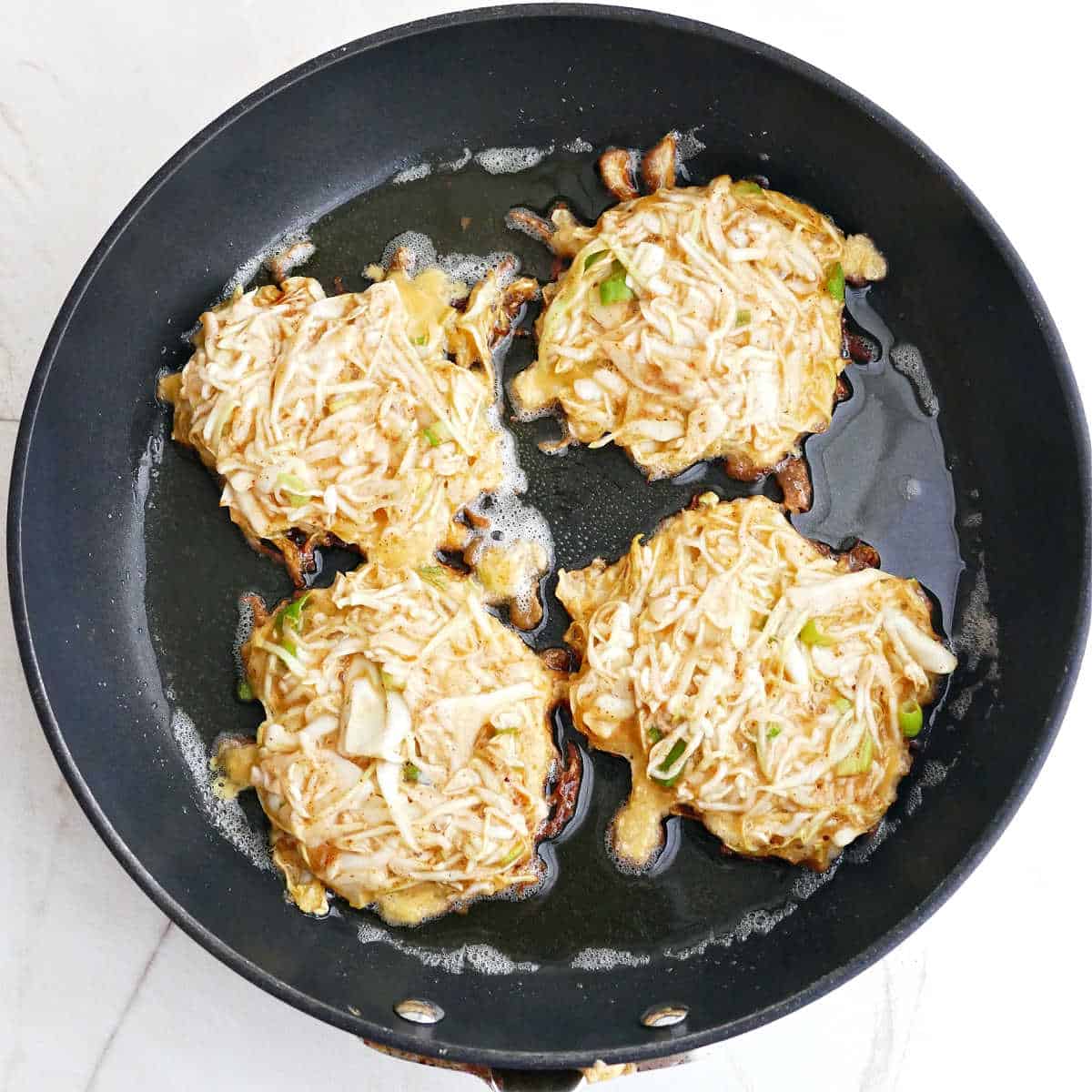 Four cabbage fritters cooking in oil in a skillet.
