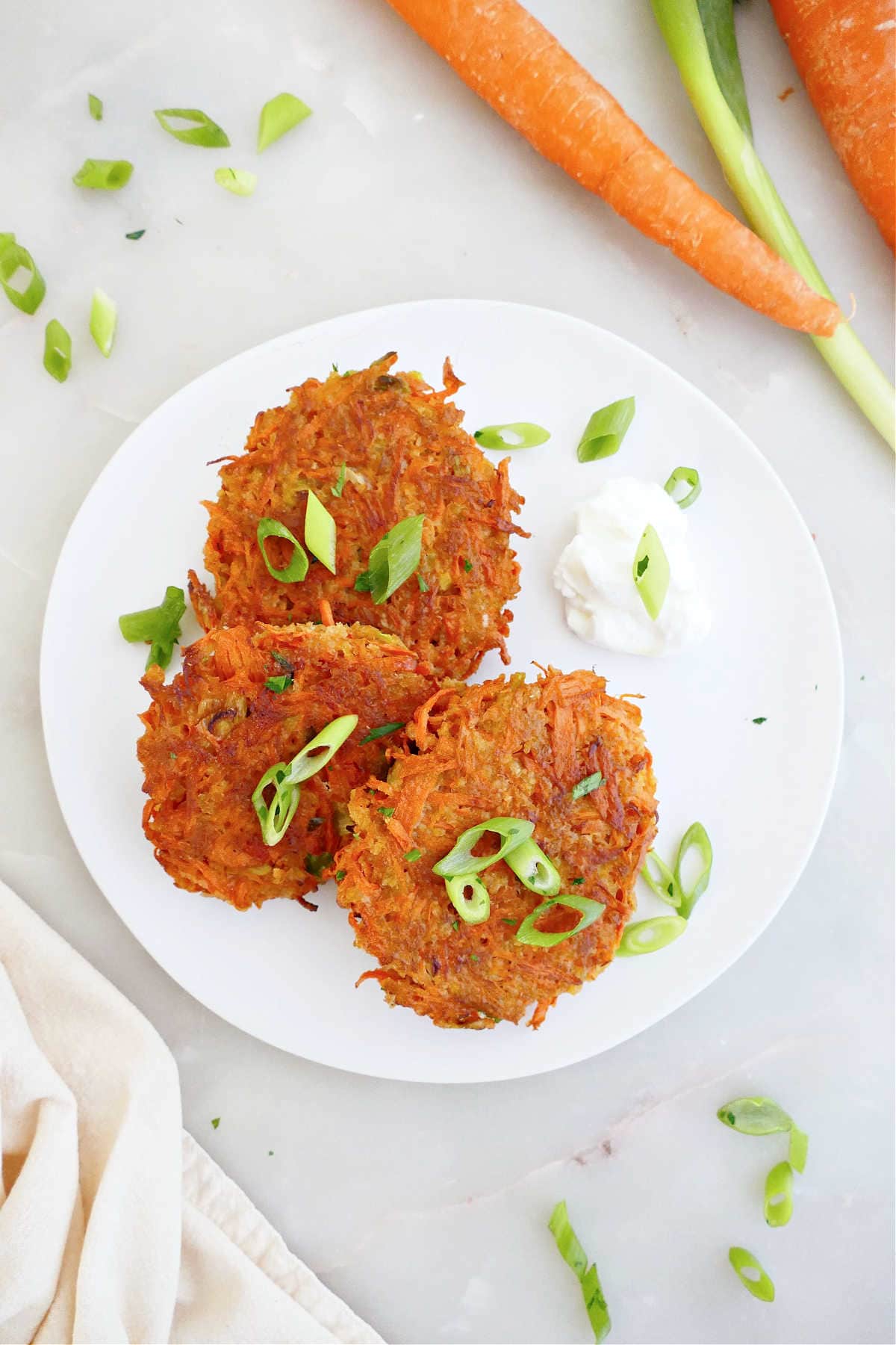 Three carrot fritters on a plate with dipping sauce and surrounded by ingredients and a napkin.