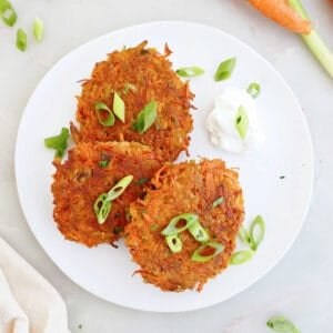 Three carrot fritters on a plate with dipping sauce and surrounded by ingredients and a napkin.