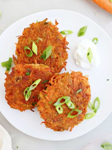 Three carrot fritters on a plate with dipping sauce and surrounded by ingredients and a napkin.