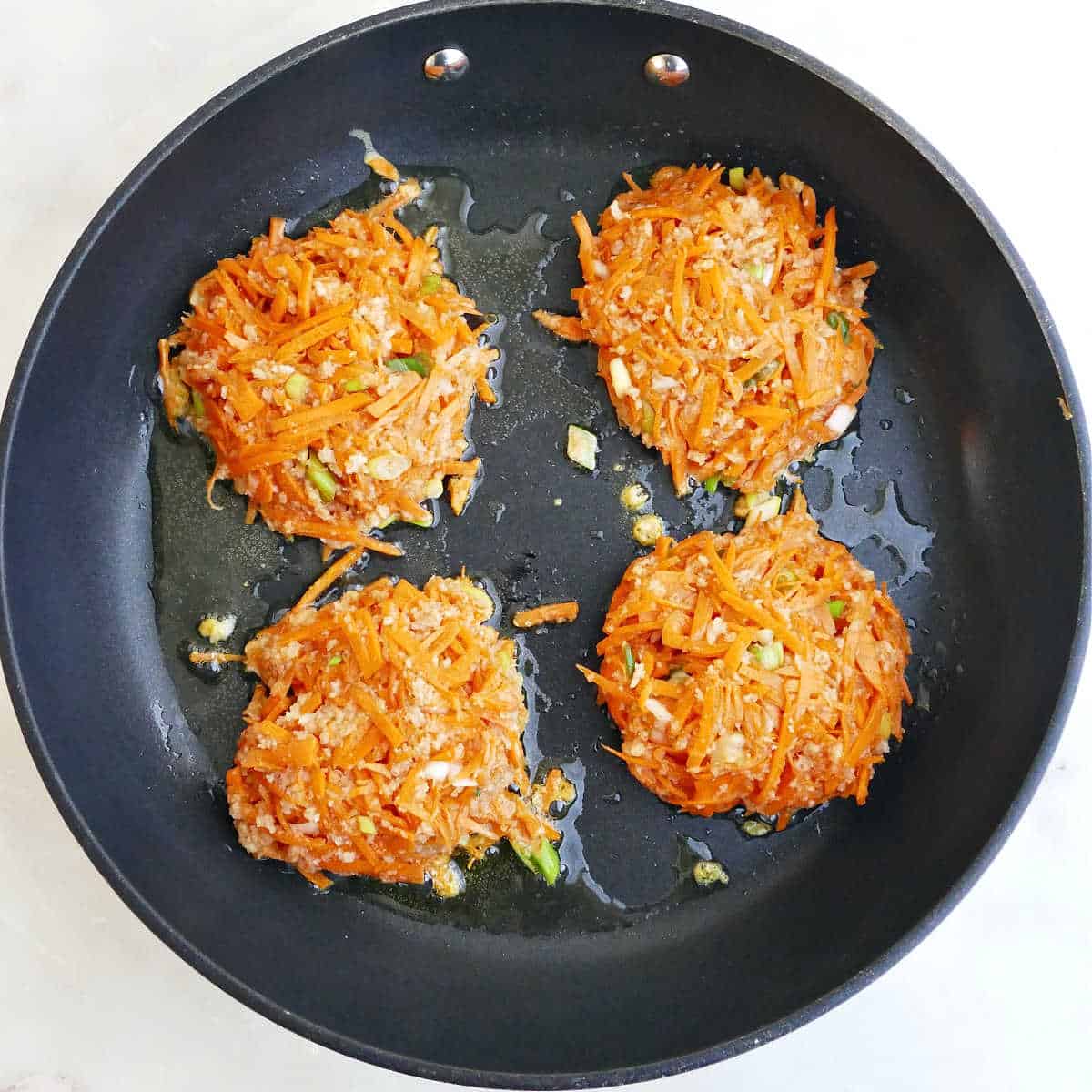 Four carrot fritters cooking in oil in a nonstick skillet.