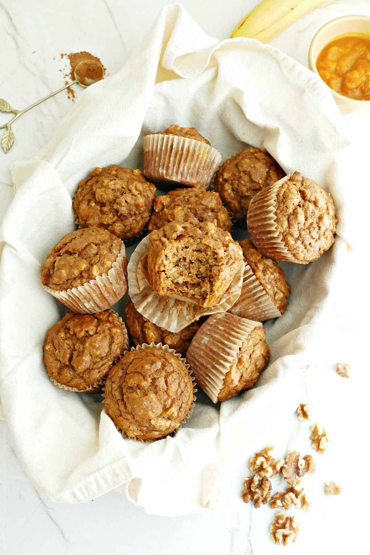 Pumpkin banana muffins in a basket lined with a napkin surrounded by ingredients.