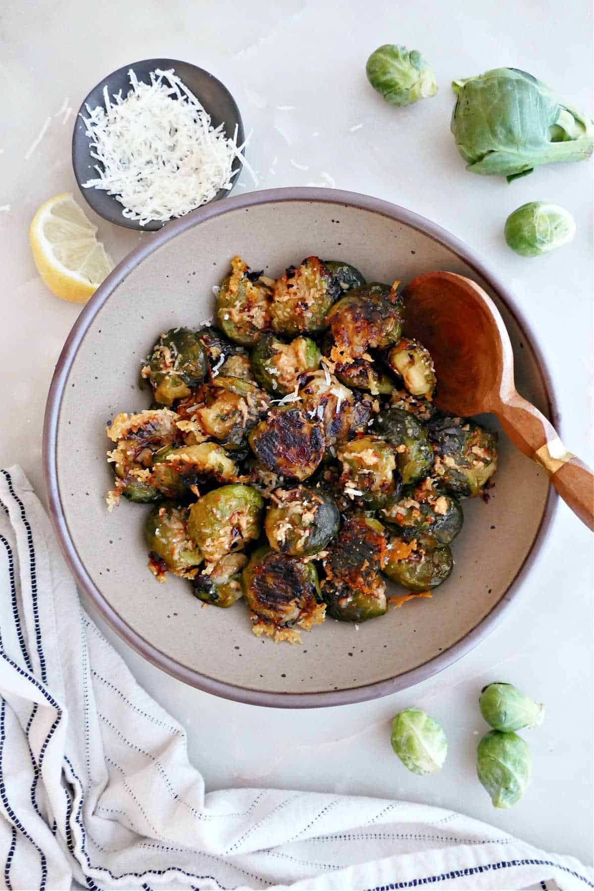 Smashed Brussels sprouts in a bowl with a wooden spoon surrounded by ingredients.