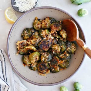 Smashed Brussels sprouts in a bowl with a wooden spoon surrounded by ingredients.