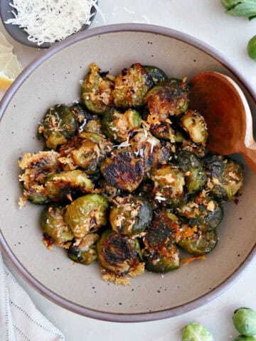 Smashed Brussels sprouts in a bowl with a wooden spoon surrounded by ingredients.