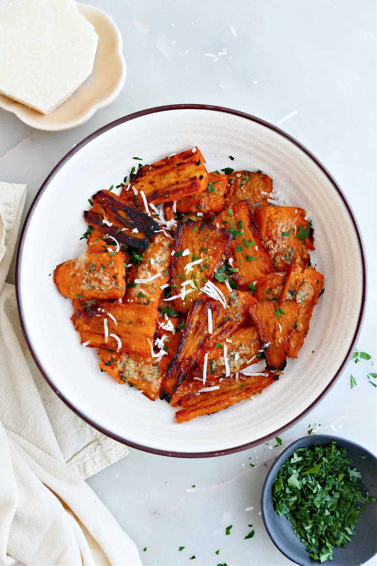 Parmesan smashed carrots in a bowl next to parsley, cheese, and a napkin.