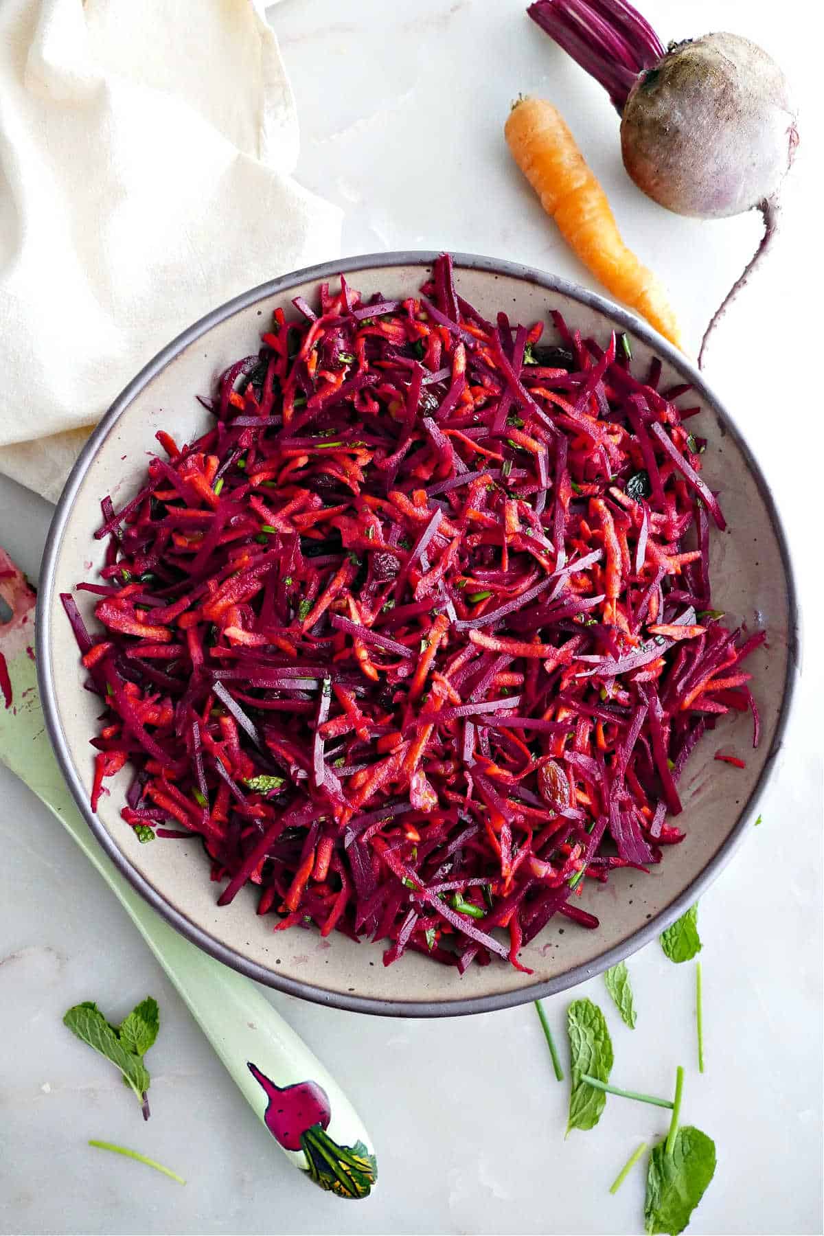 Carrot beet salad in a serving bowl next to ingredients, spoon, and napkin.