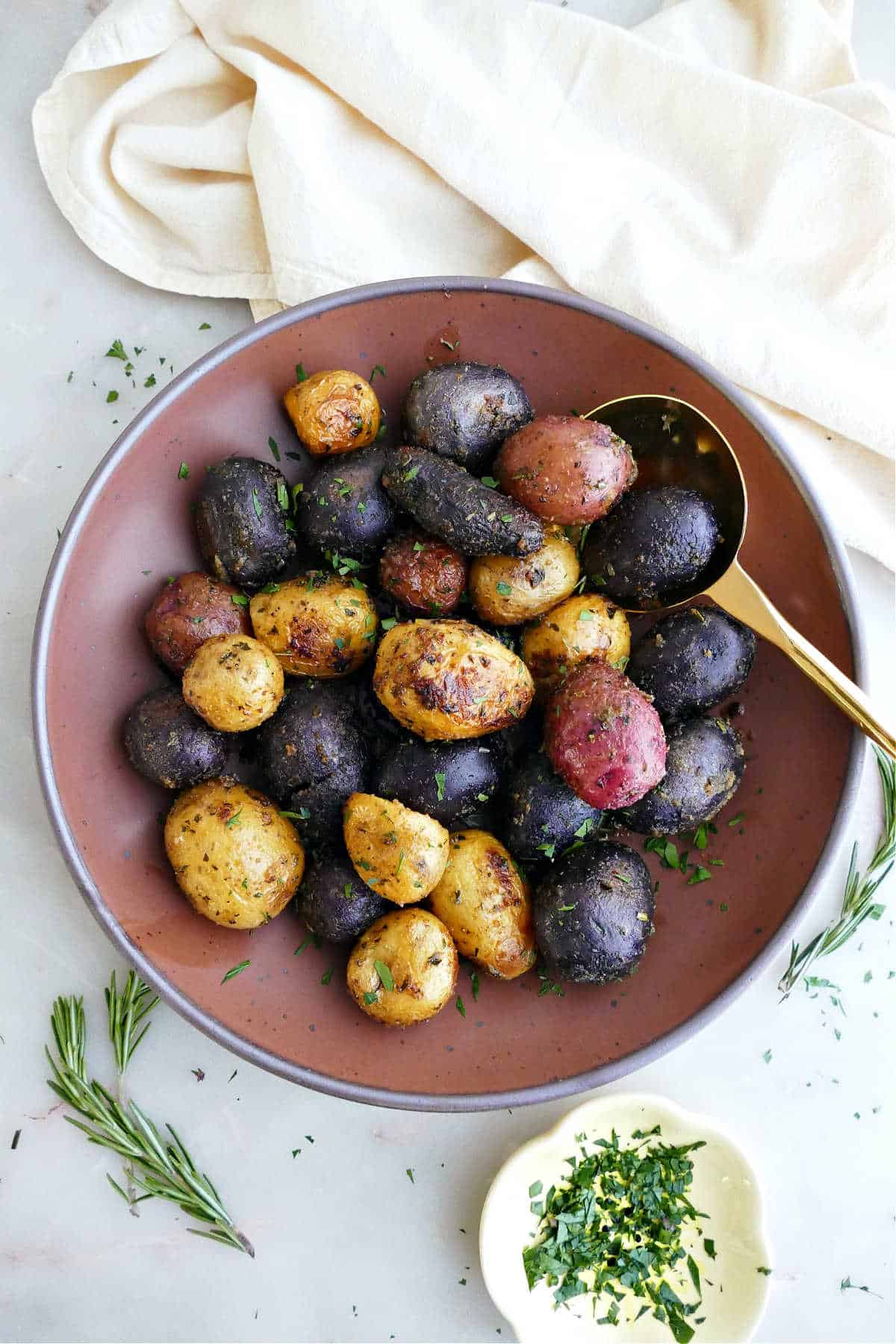 Roasted potatoes in a serving bowl with a spoon next to herbs and a napkin.